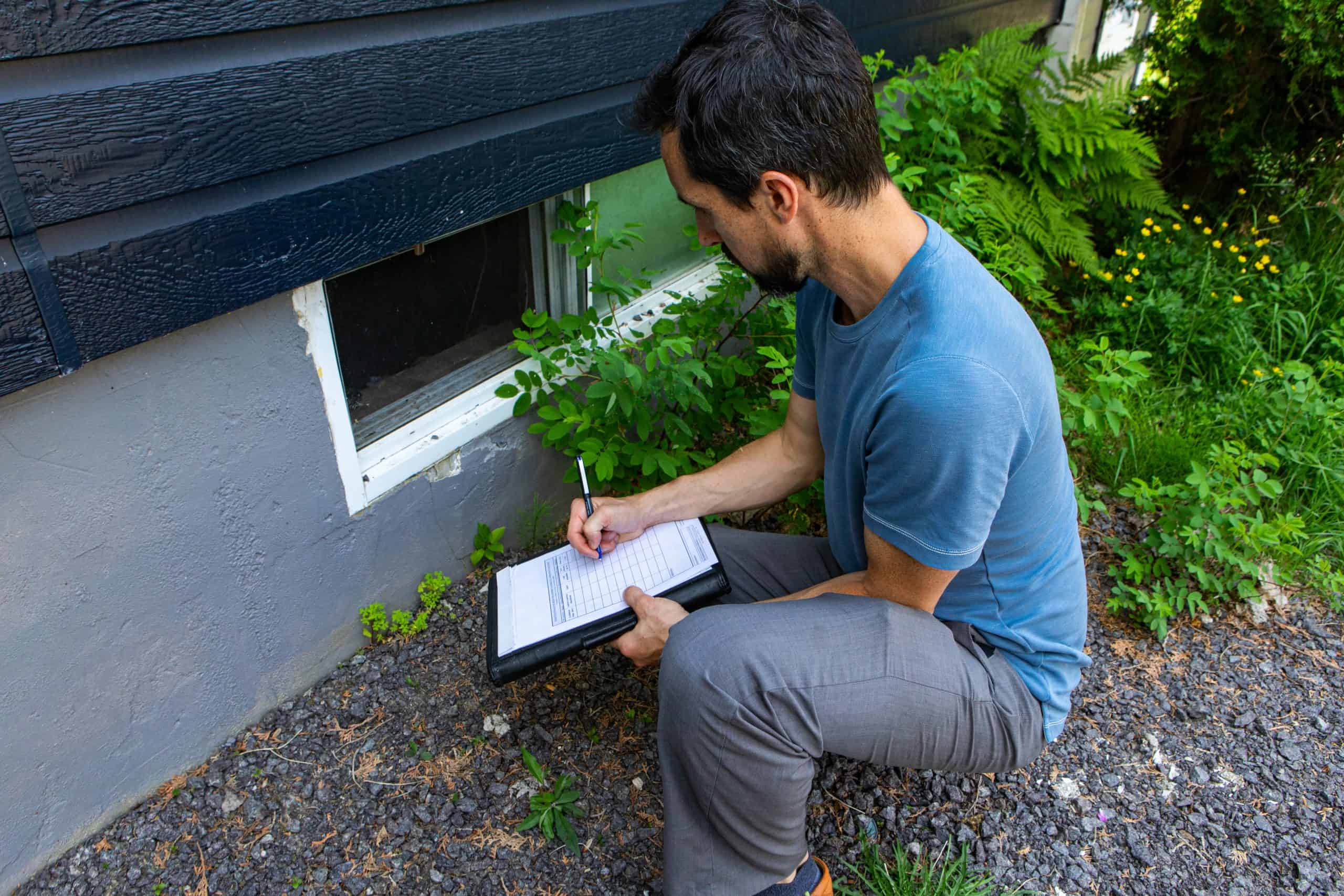 A hardscaping contractor kneels on gravel beside a house with dark siding, holding a clipboard and pen. He seems to be inspecting or taking notes near a basement window. Green plants and foliage add a touch of nature to the scenic area.