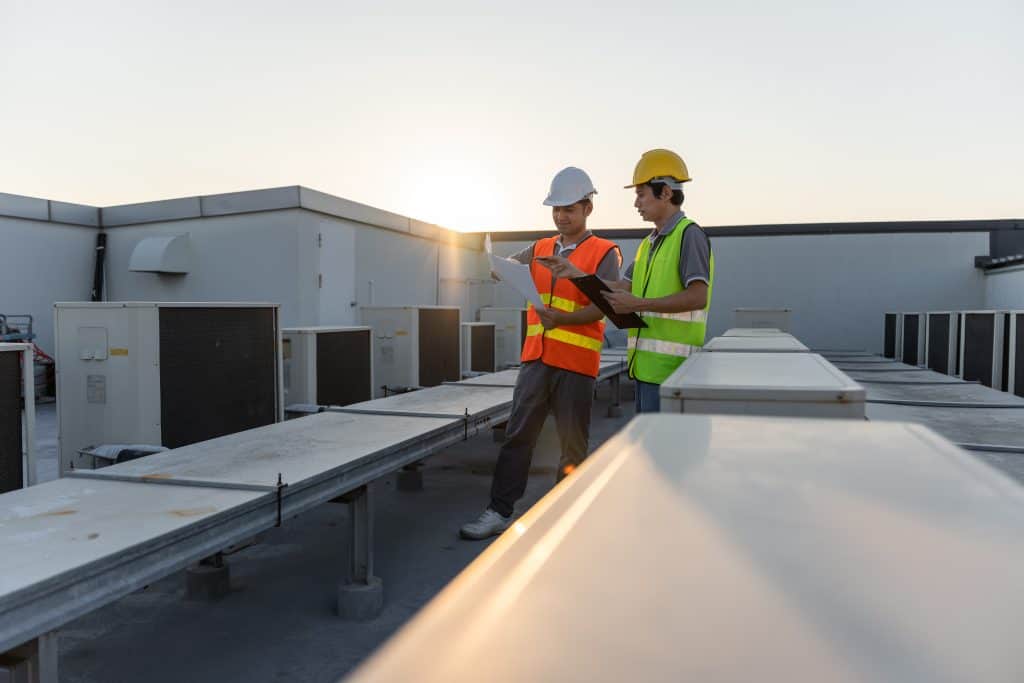 Two construction workers wearing safety gear and hard hats are standing on a rooftop next to large HVAC units. One, possibly a hardscaping contractor, is holding a clipboard, and they appear to be discussing the equipment as the sun sets in the background.