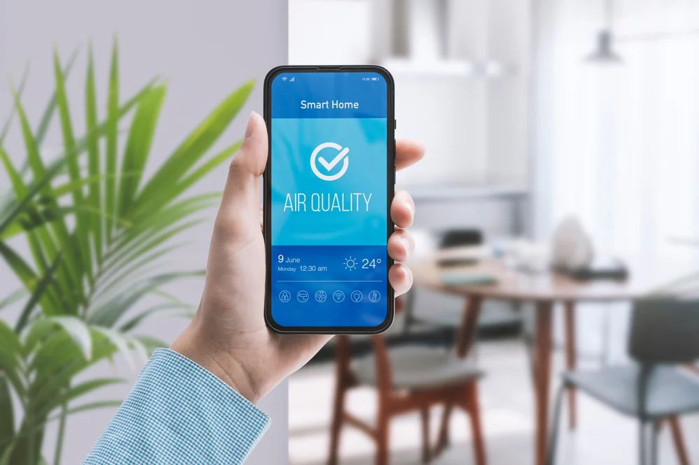 Hand holding smartphone displaying smart home air quality app with indoor air status check, modern living room background and connected home technology.