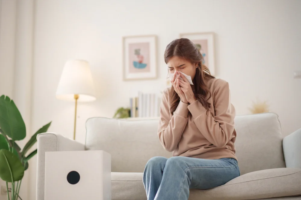 Person sitting on a sofa beside a home air purifier, indicating indoor air quality concerns, allergy relief, or air filtration use in a modern living room.